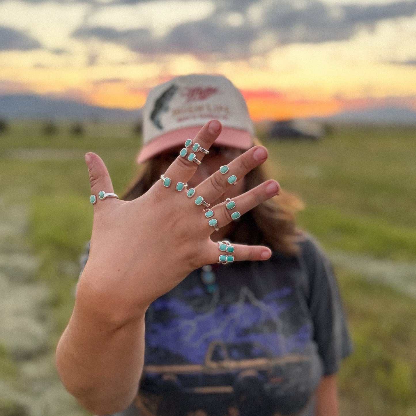 Person in a field with a sunset, wearing multiple turquoise rings on their hand.