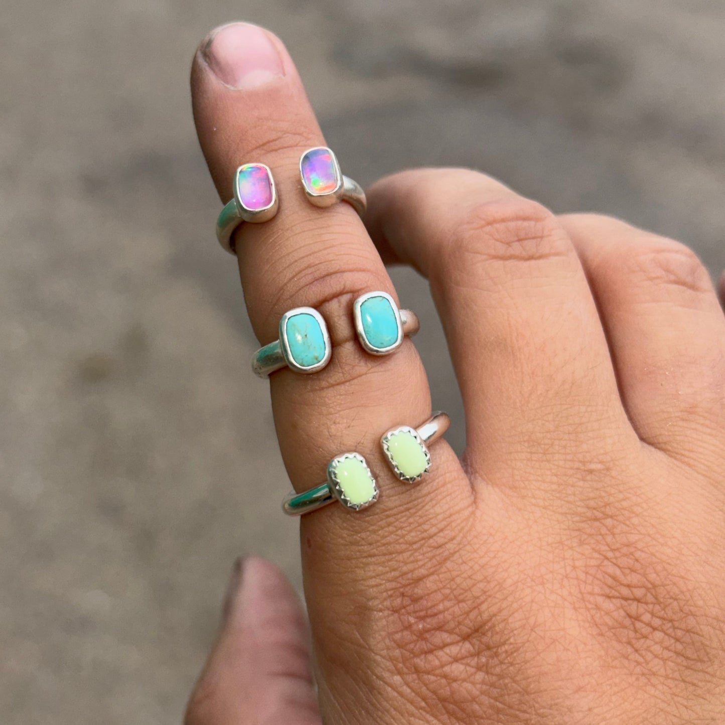 A close-up of a hand wearing three silver rings with gemstones against a blurred gray concrete background.